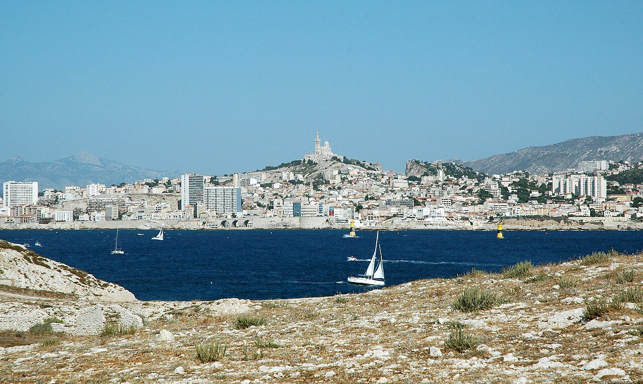 Marseille vue depuis l’île Ratonneau : la rade bleue et un voilier, la ville blanche au pied de Notre-Dame de la Garde, les collines au fond