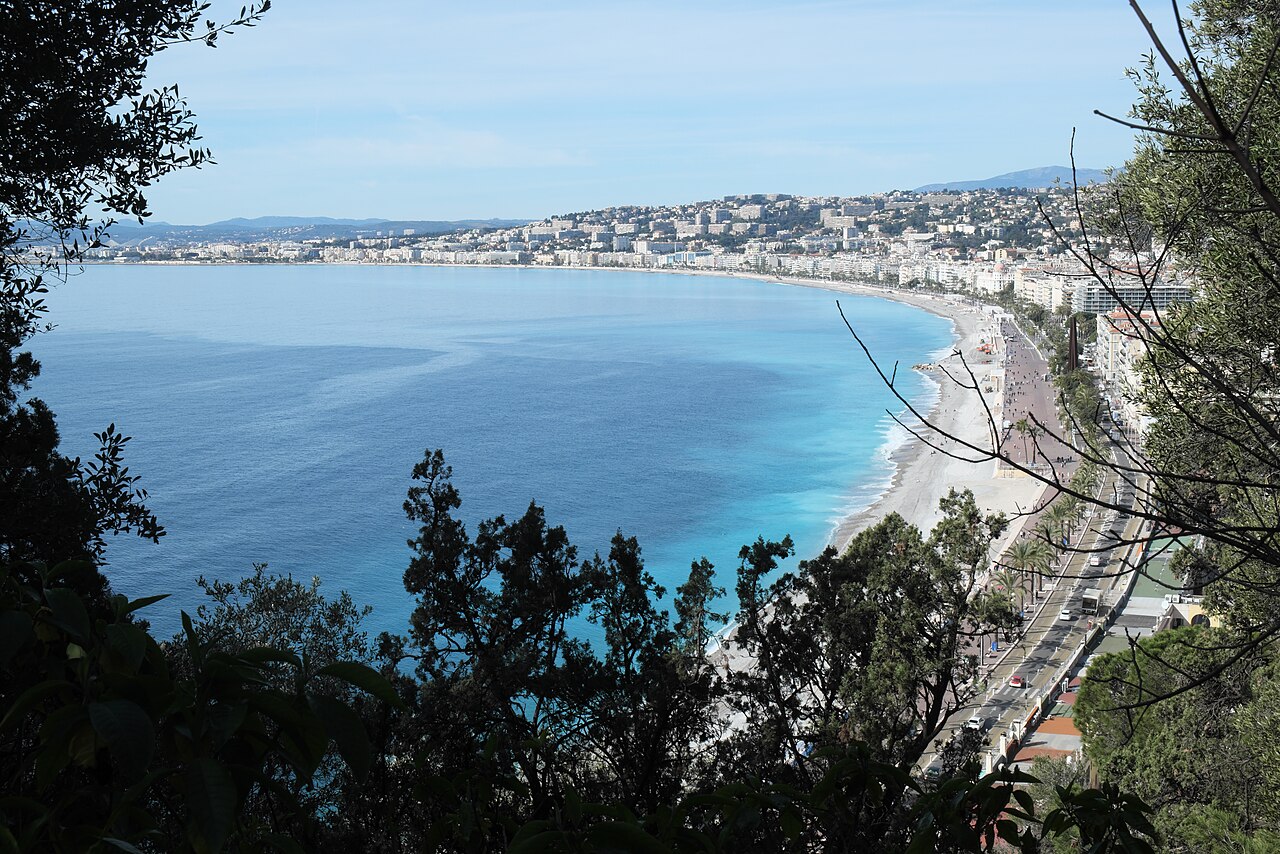 La courbe de la baie des Anges et de la promenade des Anglais vue d’en haut, entre les arbres de la colline du Château