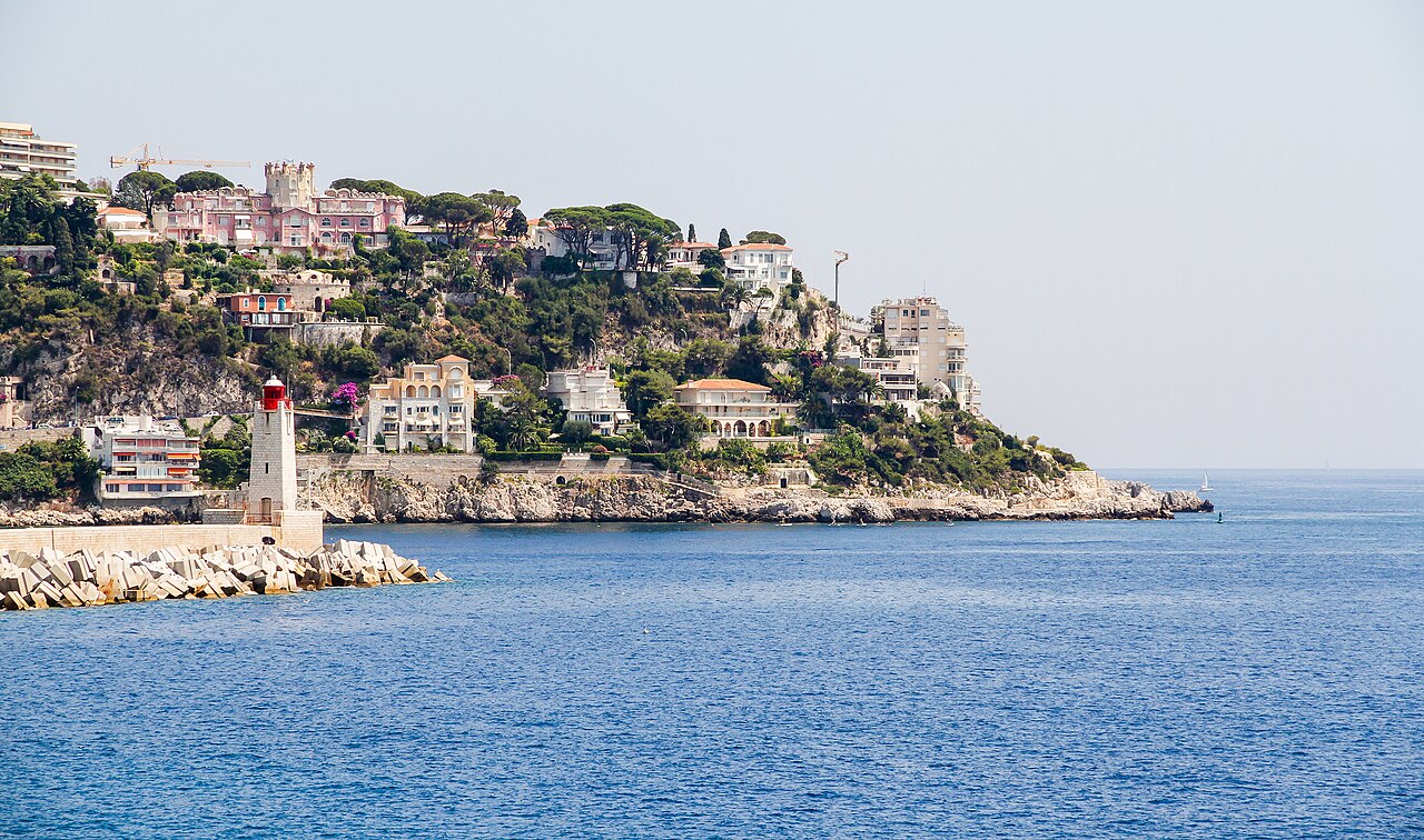Le cap de Nice et ses villas au-dessus des rochers, le phare rouge et blanc de la jetée du port et la mer bleue