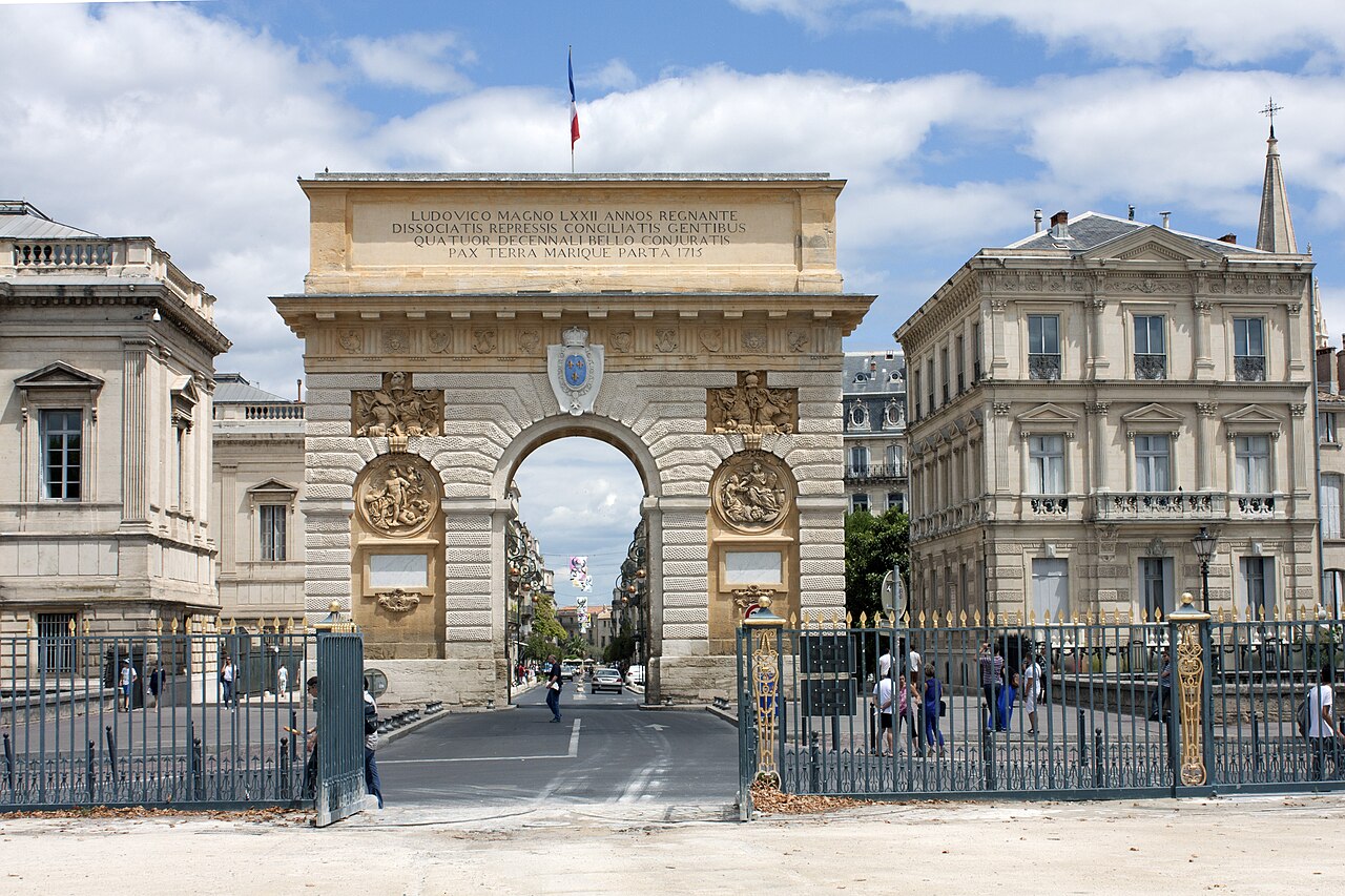 L’arc de triomphe de Montpellier (porte du Peyrou), en pierre blonde sculptée, vu depuis les grilles de la promenade du Peyrou, la rue Foch en enfilade sous l’arche