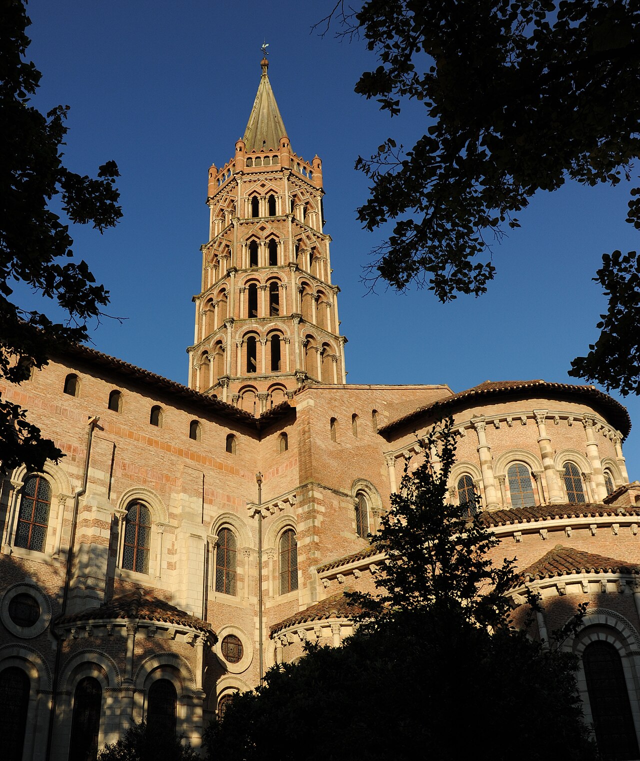 Le chevet roman de brique et de pierre de la basilique Saint-Sernin de Toulouse, ses chapelles rayonnantes et son clocher octogonal à étages, encadrés de feuillages sous un ciel bleu