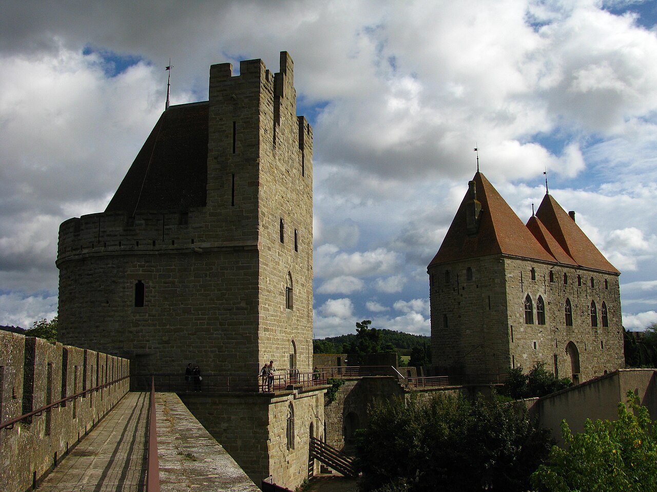 Le chemin de ronde des remparts nord de la Cité de Carcassonne, une grosse tour crénelée et une tour-porte aux toits de tuiles, sous un ciel nuageux