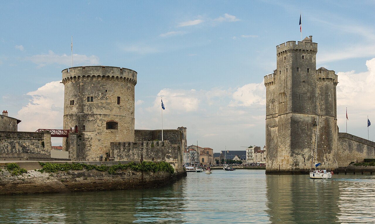 L’entrée du Vieux-Port de La Rochelle vue depuis l’eau, entre la tour ronde de la Chaîne et la tour Saint-Nicolas, avec un voilier et les quais au fond