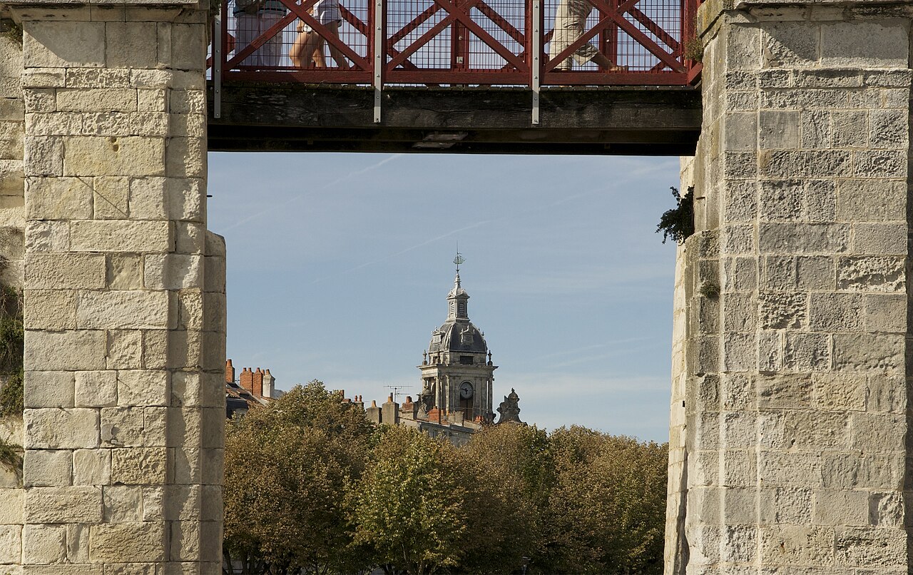 Le campanile à dôme et à horloge de la Grosse Horloge de La Rochelle, vu au-dessus des arbres, encadré par deux piliers de pierre sous une passerelle