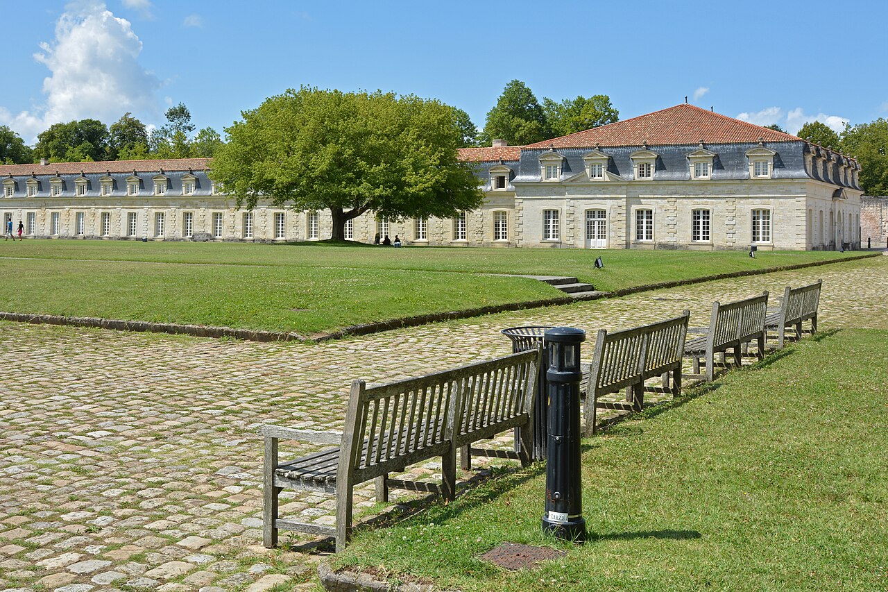 La longue façade classique de la Corderie royale de Rochefort, derrière sa pelouse, un grand arbre et une allée pavée bordée de bancs