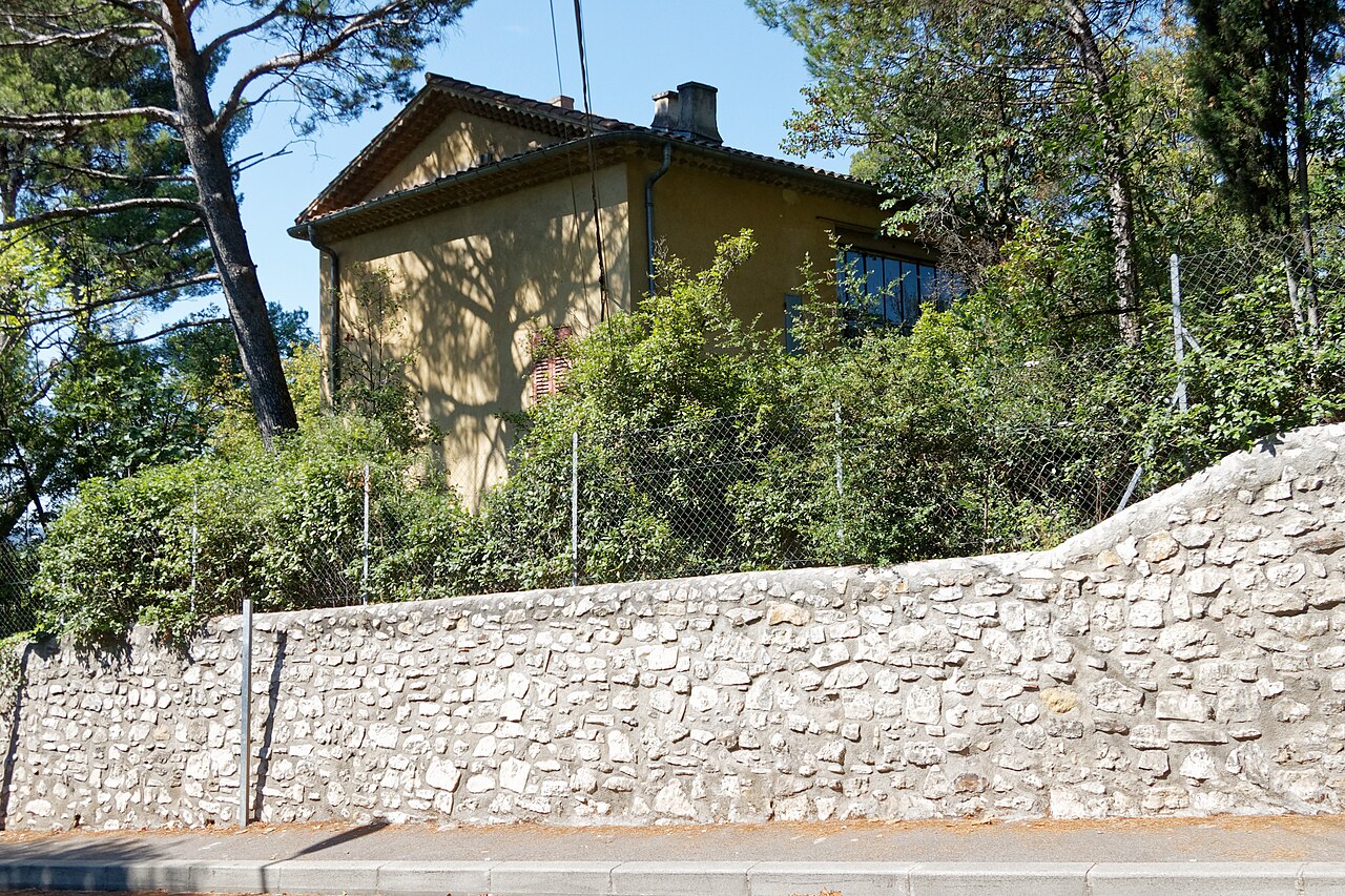La maison-atelier de Cézanne aux Lauves, ocre et coiffée de tuiles, au-dessus d’un mur de pierres sèches et d’un jardin touffu, sous un ciel bleu