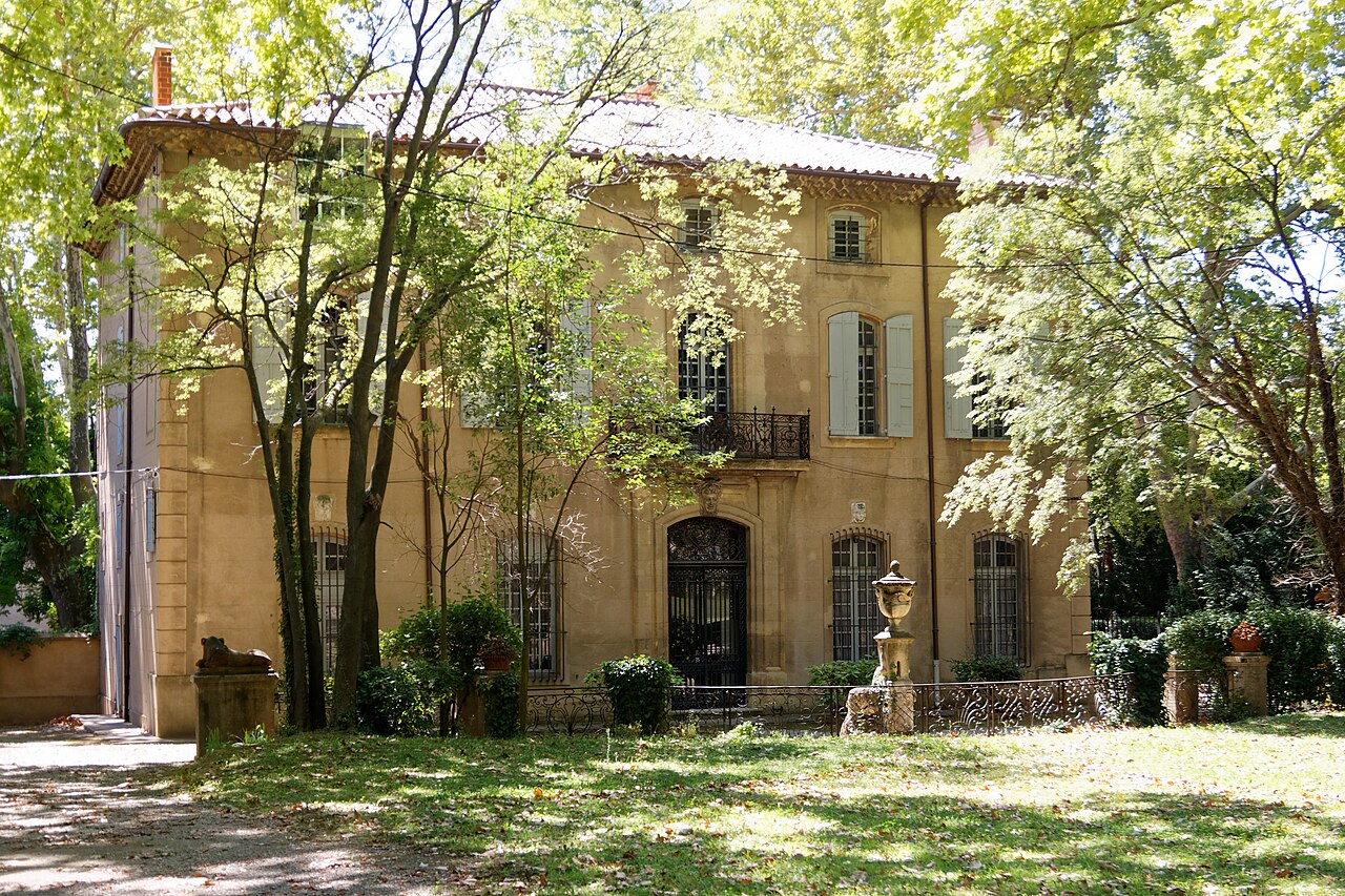 La façade ocre de la bastide du Jas de Bouffan, ses volets gris et son balcon en fer forgé, derrière les arbres du parc