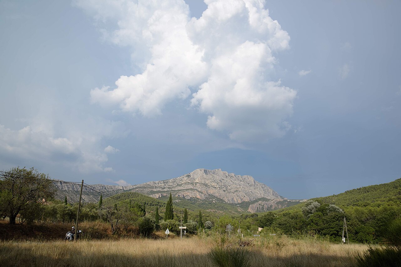 La montagne Sainte-Victoire et sa crête calcaire sous un grand nuage d’orage, au-dessus de la garrigue, de cyprès et d’un champ d’herbes sèches