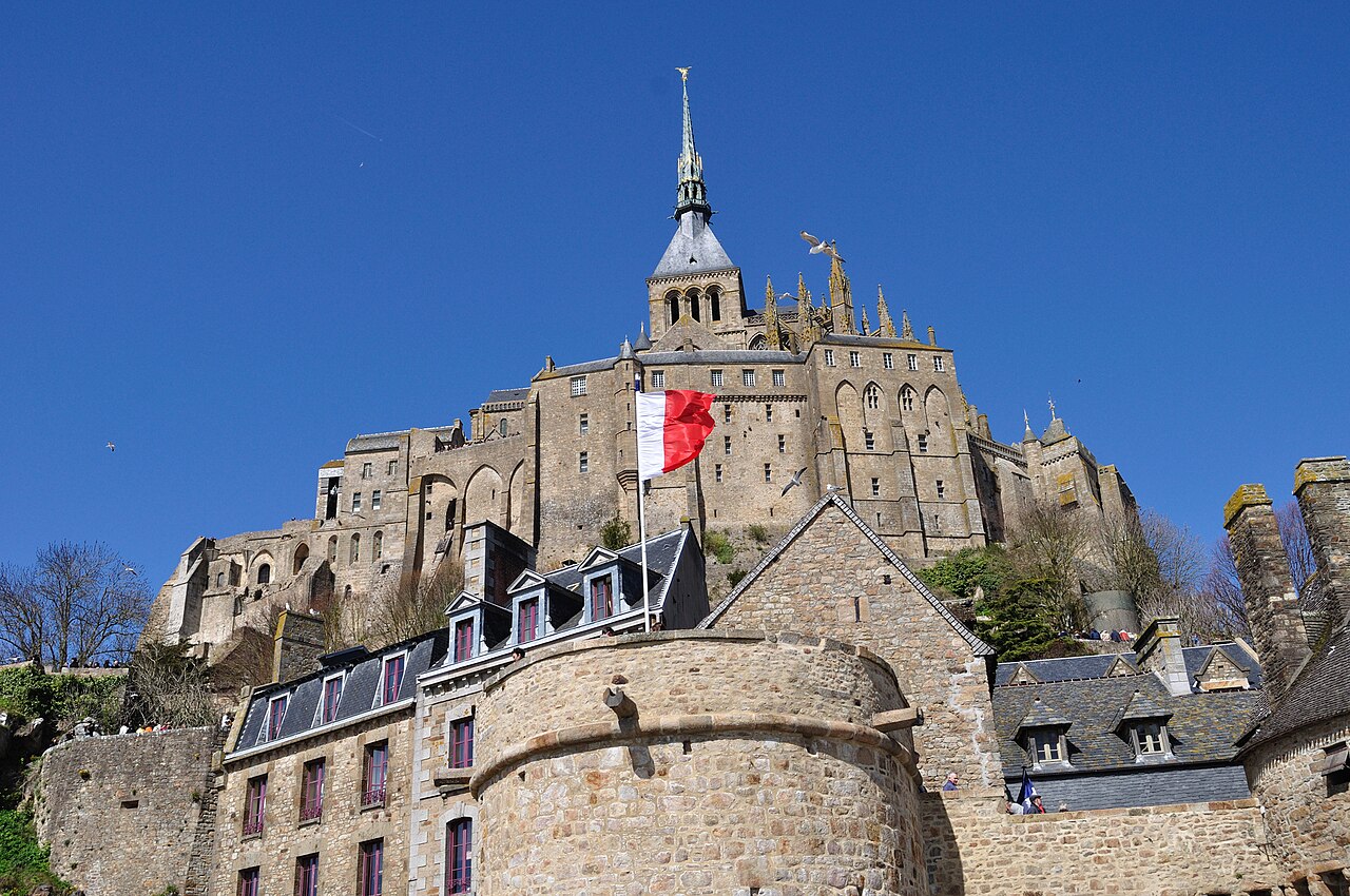 L’abbaye du Mont-Saint-Michel et sa flèche dorée vues d’en bas, au-dessus des remparts et des maisons de granit du village, sous un ciel bleu
