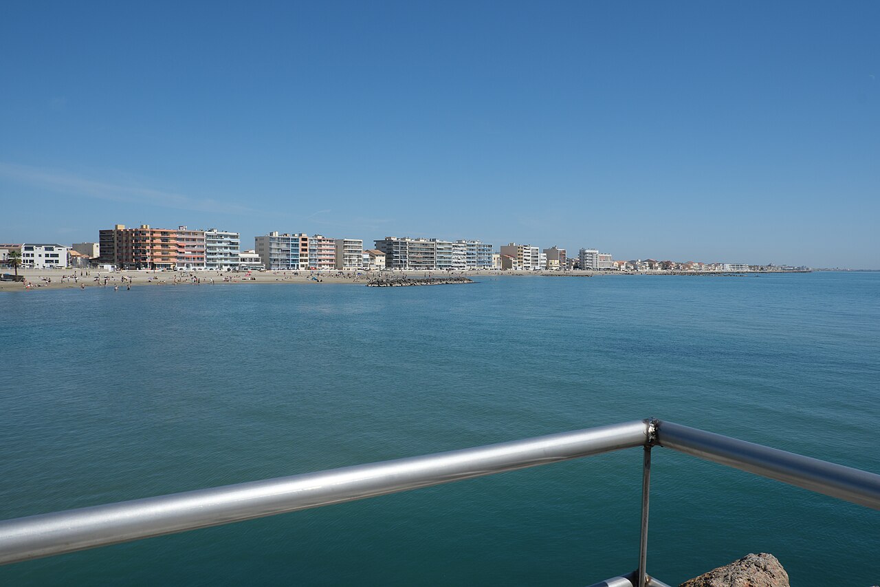 La plage de sable de Palavas-les-Flots et sa mer calme et turquoise, vues depuis une jetée, avec la ligne des immeubles du front de mer sous un ciel bleu