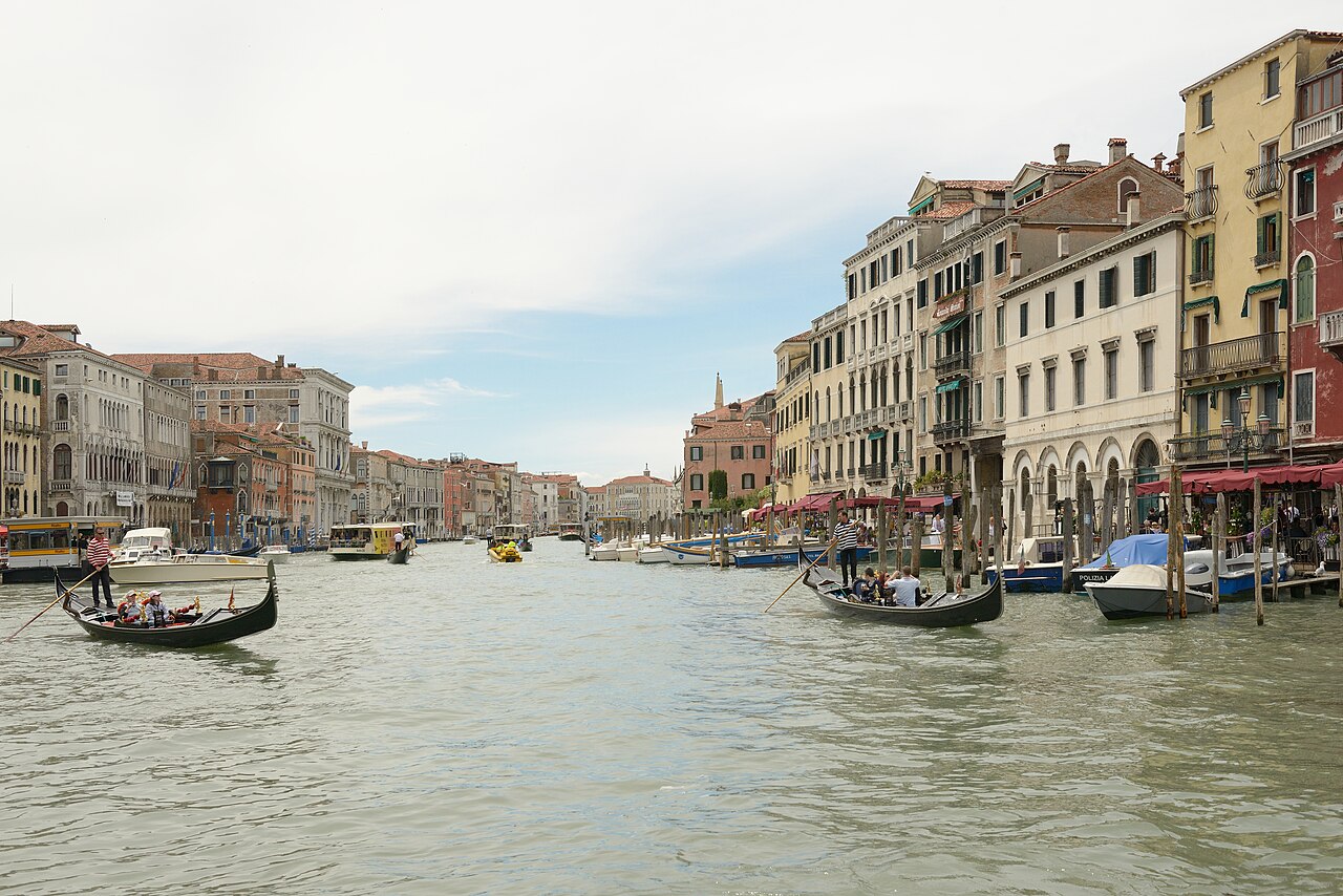 Le Grand Canal à Venise vu du pont du Rialto : deux gondoles sur l’eau verte, les palais aux façades ocre et rouges de la Riva del Vin à droite, des vaporetti au loin