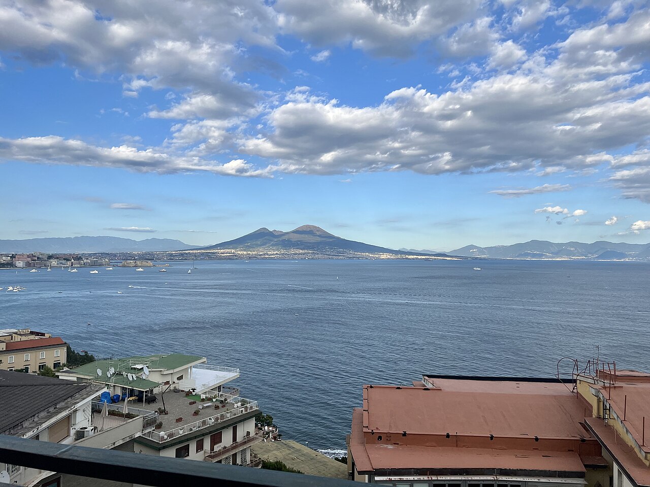 Le golfe de Naples vu de la colline du Pausilippe : la mer bleue parsemée de bateaux, la ville au loin et le Vésuve sous un ciel de nuages, des toits en terrasse au premier plan