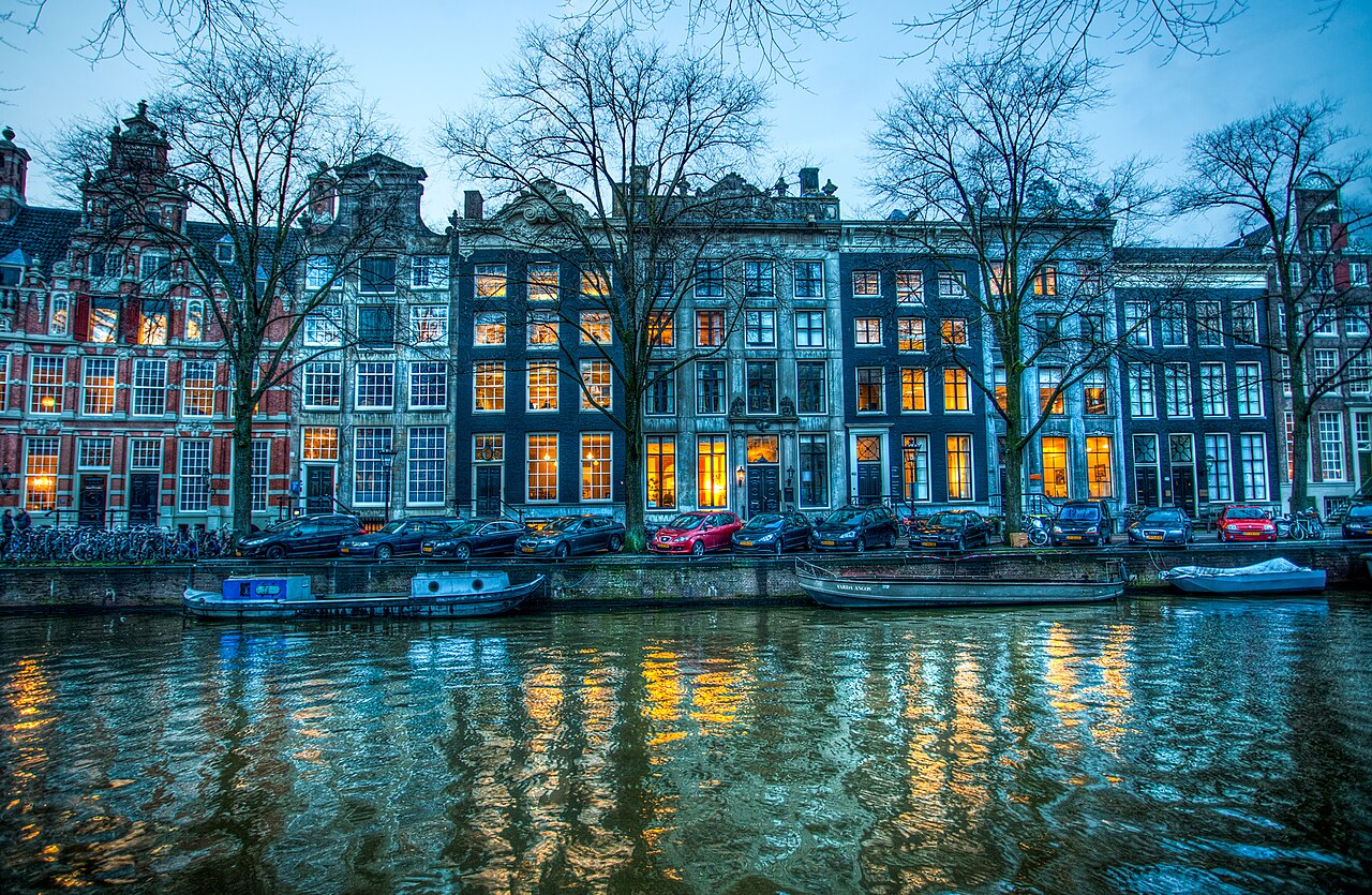 Les maisons à pignon du Herengracht à Amsterdam à l’heure bleue, fenêtres éclairées se reflétant dans le canal, arbres nus et péniches le long du quai