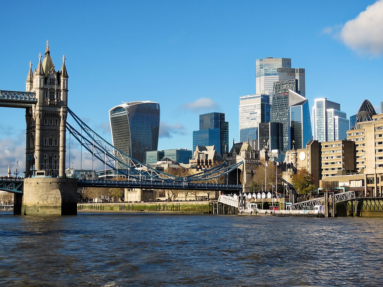 Tower Bridge et les tours de la City de Londres vus depuis la Tamise, sous un ciel bleu, la tour de Londres et ses quais au pied des gratte-ciel