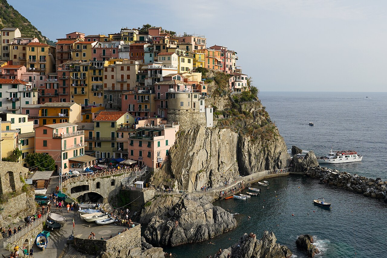 Le village de Manarola, dans les Cinque Terre, et ses maisons colorées perchées sur un éperon rocheux au-dessus d’un petit port, un bateau de passagers en mer
