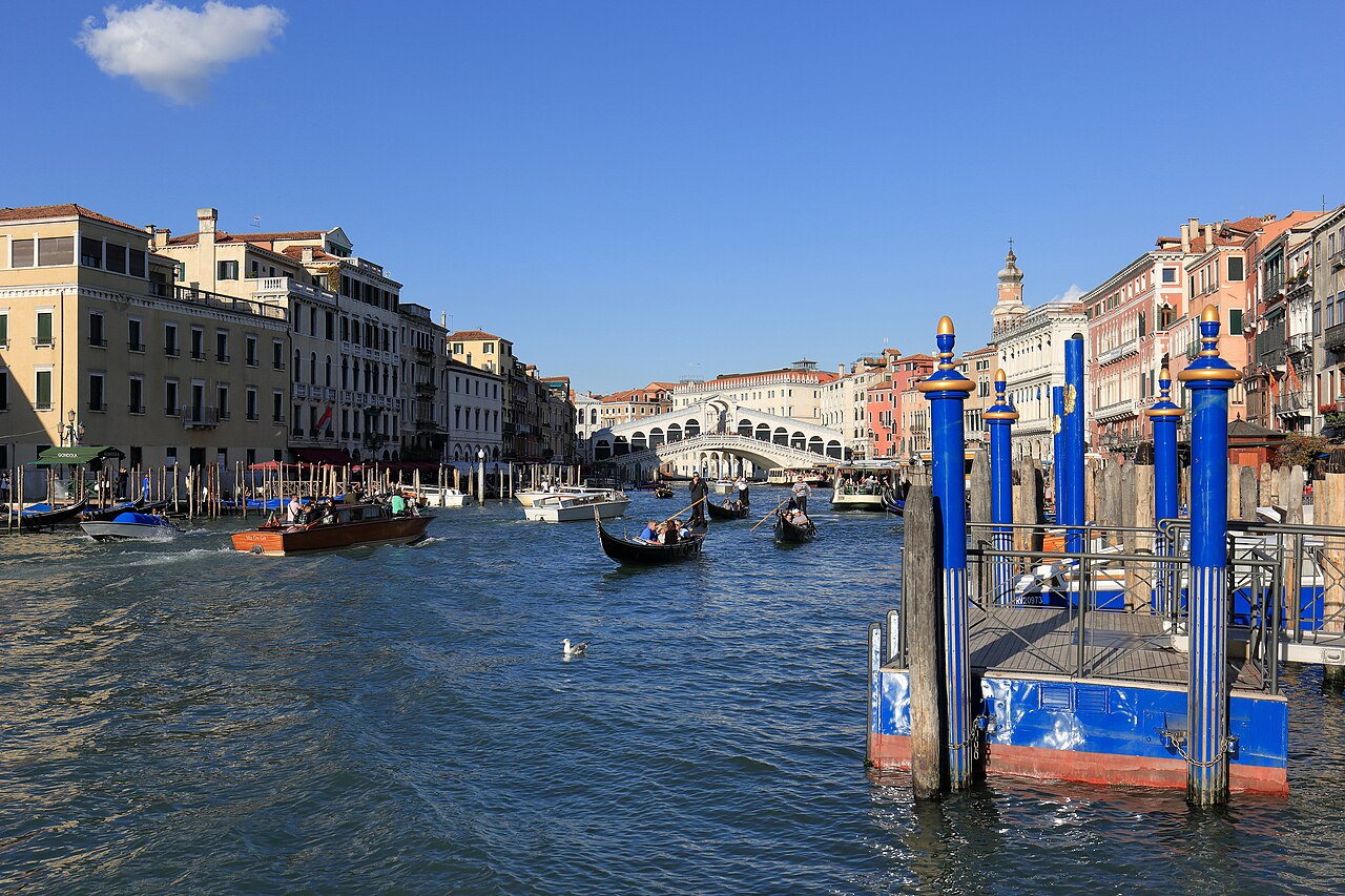 Le Grand Canal par beau temps, des gondoles et des bateaux-taxis sur l’eau, le pont du Rialto au fond et les poteaux d’amarrage bleus d’un ponton à droite