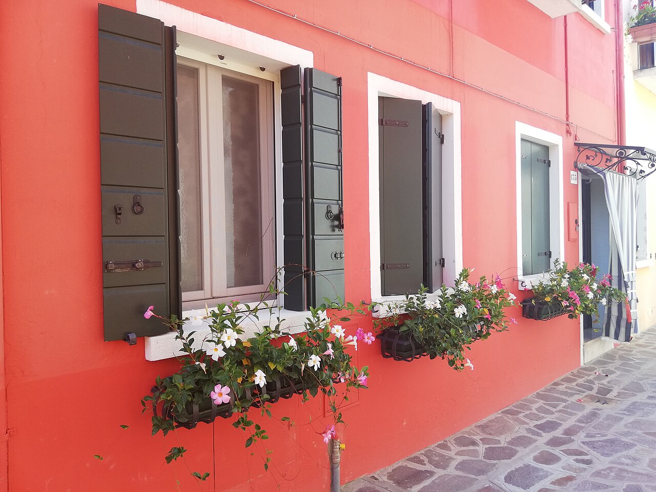 Une maison de Burano peinte en rouge corail, ses volets verts et ses jardinières de fleurs, au bord d’une ruelle pavée