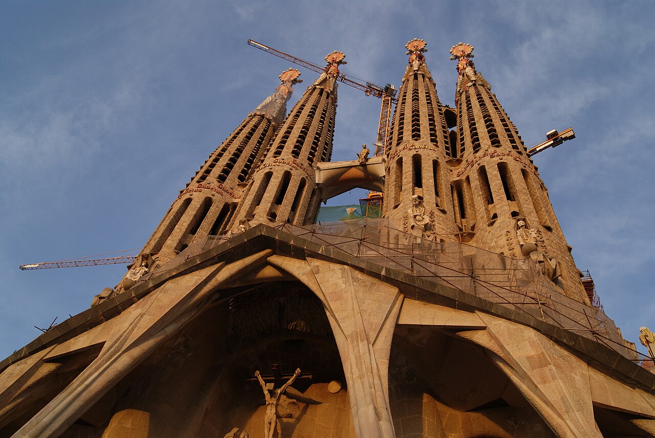 La façade de la Passion de la Sagrada Família vue d’en bas : les quatre clochers ajourés, le portique aux colonnes inclinées et des grues de chantier contre le ciel