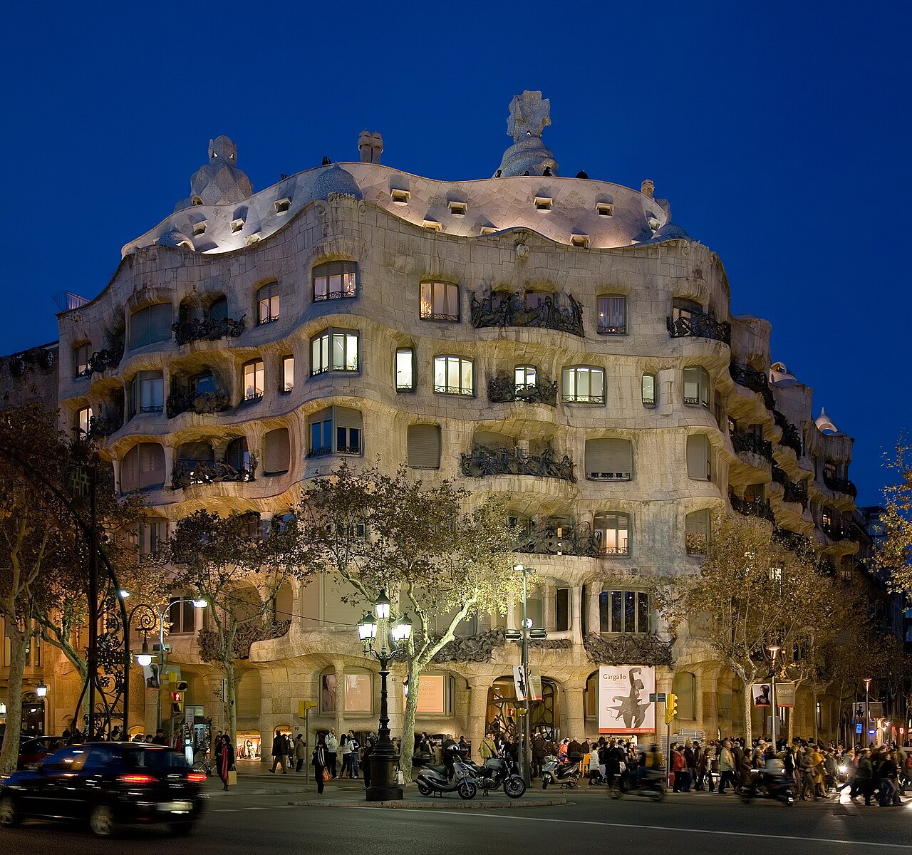 La Pedrera (Casa Milà) au crépuscule, à l’angle du passeig de Gràcia : la façade de pierre ondulée et ses balcons de fer forgé éclairés
