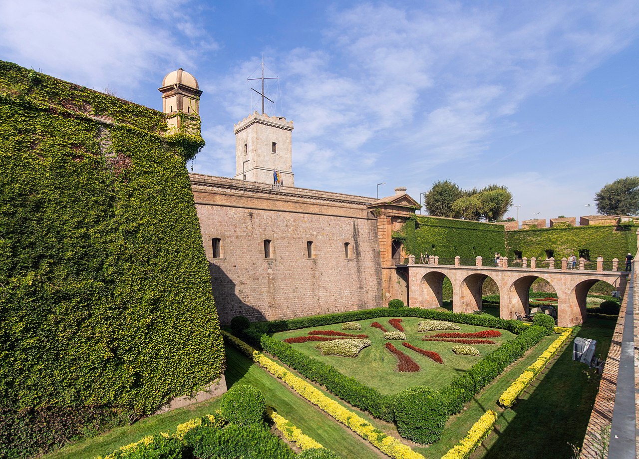 Le château de Montjuïc : remparts couverts de lierre, fossé aménagé en jardin et pont d’accès sous un ciel bleu