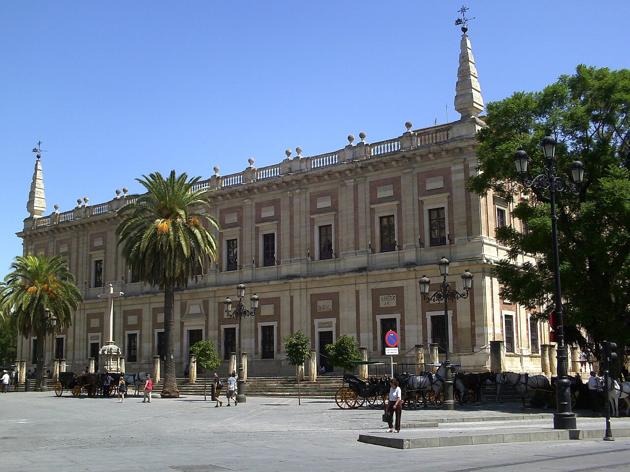 L’Archivo General de Indias, édifice Renaissance à balustrade, avec des palmiers et des calèches sur la place