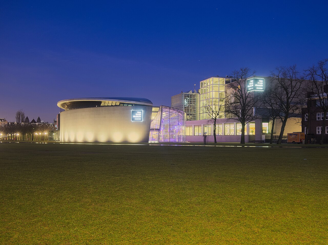 Le musée Van Gogh à l’aube, aile ovale et bâtiment vitré éclairés, vus depuis la pelouse du Museumplein